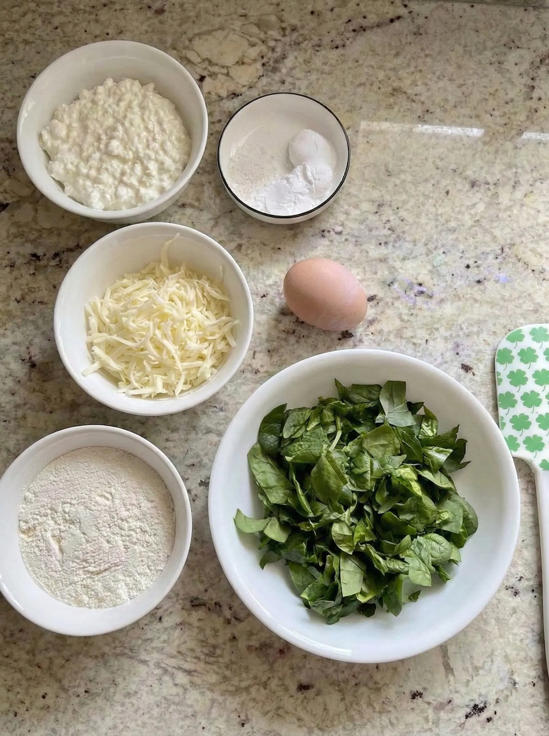 Overhead view of the simple ingredients needed for spinach cottage cheese flagels arranged on a speckled countertop. Bowls contain cottage cheese, all-purpose flour, shredded mozzarella, fresh spinach, and baking powder, with a single brown egg in the center.