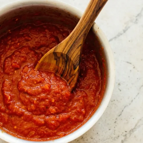 Top-down view of a white bowl filled with thick, vibrant red homemade tomato sauce made from canned tomatoes, with a rustic wooden spoon resting inside against a light marble countertop.