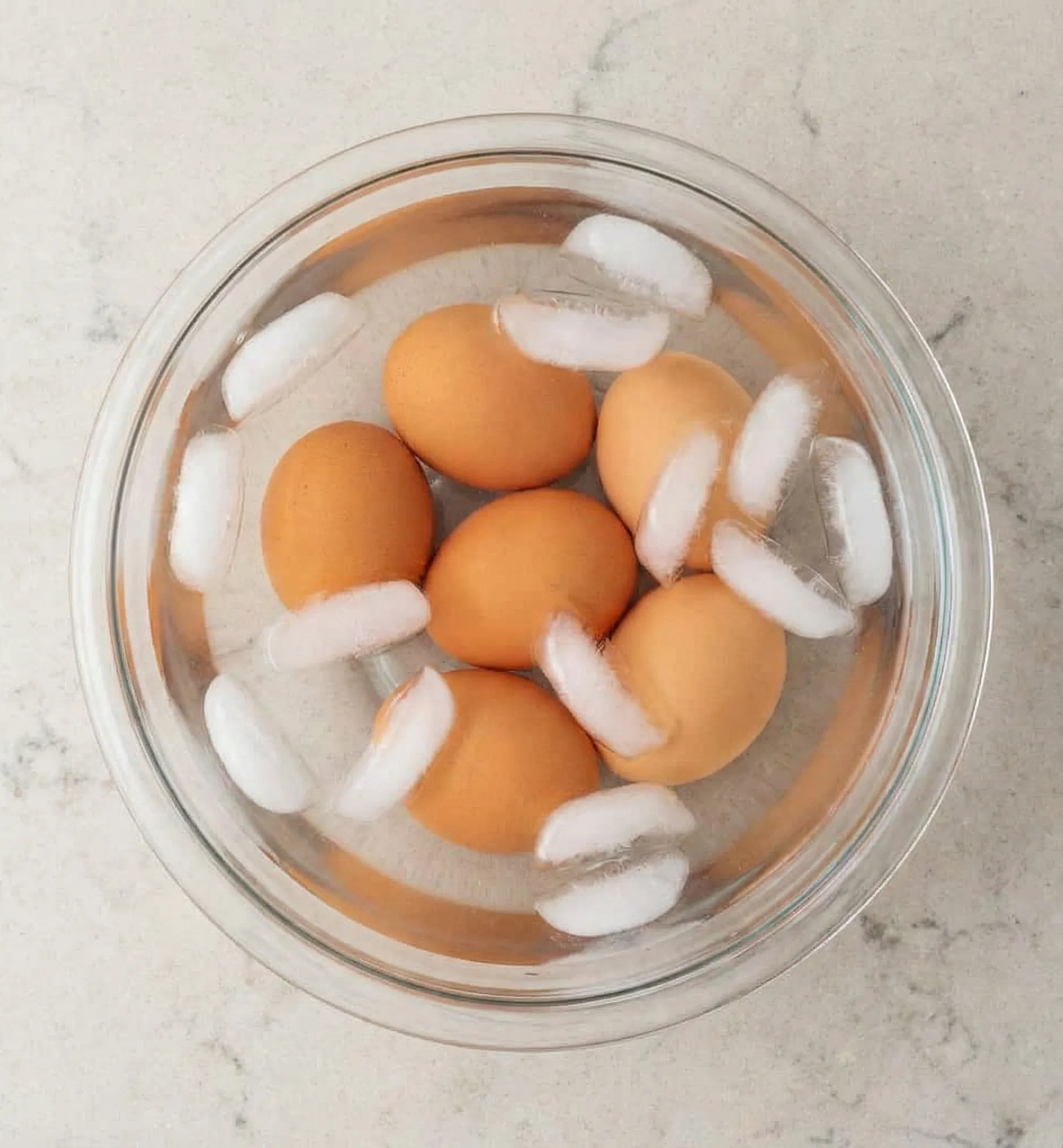 perfect hard boiled eggs A top-down photograph of a clear glass bowl filled with cold water, several ice cubes, and six brown hard-boiled eggs. The bowl is resting on a light beige countertop with grey speckles.
