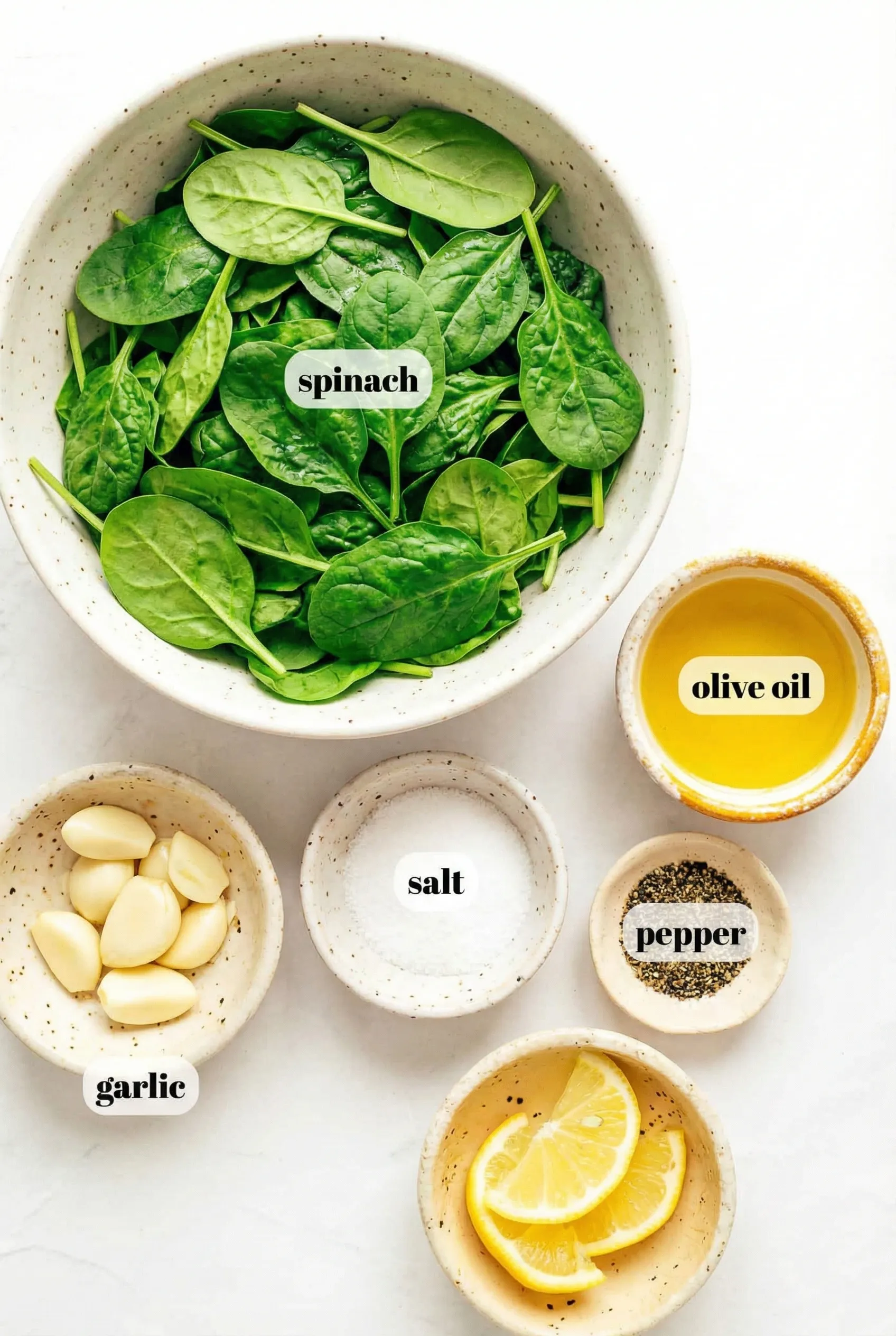 sauteed spinach ingredients A flat lay arrangement of ingredients for sautéed spinach on a white marble surface. A large bowl of fresh baby spinach sits next to small bowls containing olive oil, whole garlic cloves, sea salt, black pepper, and lemon slices. Each ingredient is labeled with black text on a white background.