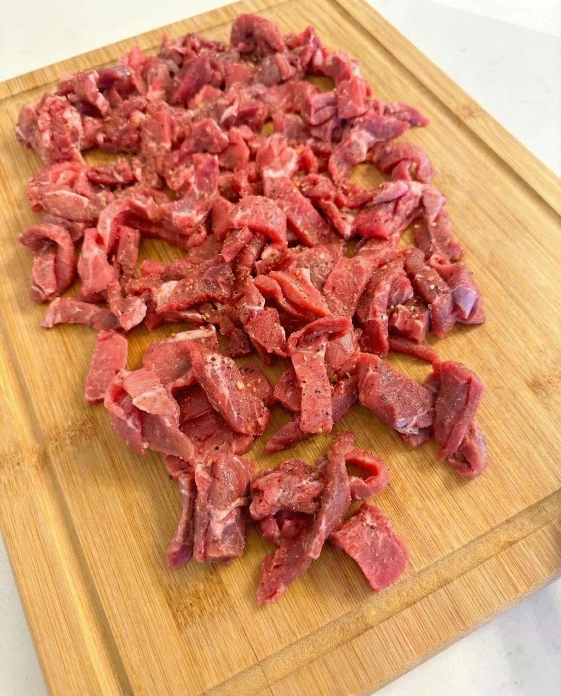 Raw sirloin steak sliced into thin strips and seasoned with spices on a wooden cutting board, ready for cooking.