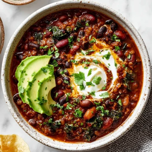 A top-down and close-up view of a high-protein bean and lentil chili bowl topped with sliced avocado, fresh cilantro, and a creamy white swirl, highlighting its rich, chunky texture.