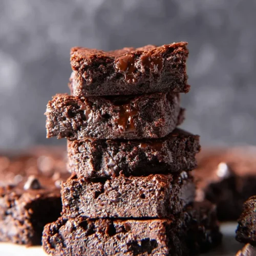 A close-up vertical stack of five super fudgy vegan and gluten-free brownies featuring visible melted chocolate chips and a dense, moist texture, surrounded by scattered chocolate chips on a surface.
