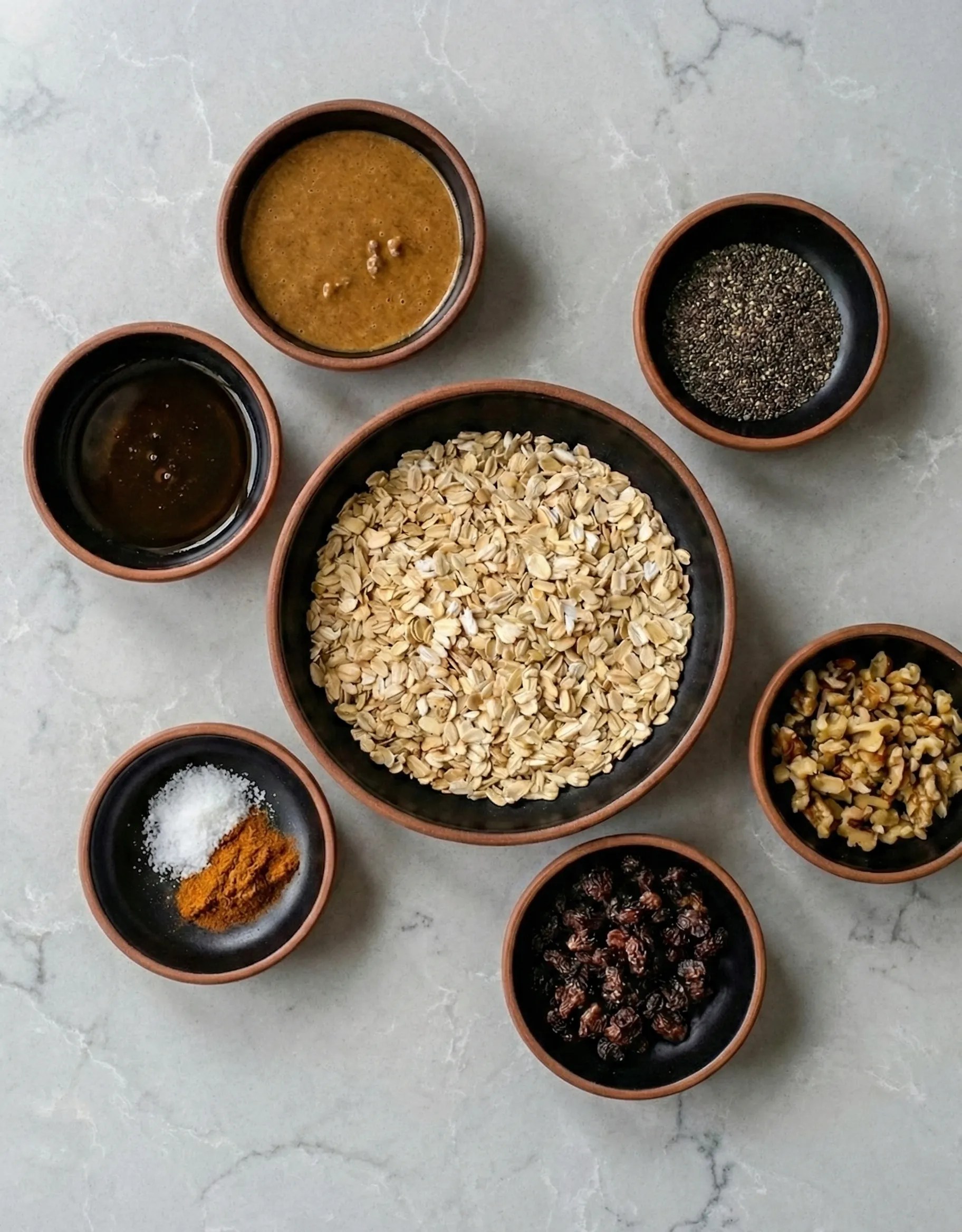 healthy energy bite ingredients Top-down view of healthy energy bite ingredients arranged in small dark bowls on a marble countertop, featuring rolled oats in the center surrounded by nut butter, chia seeds, chopped walnuts, raisins, cinnamon, salt, and liquid sweetener.