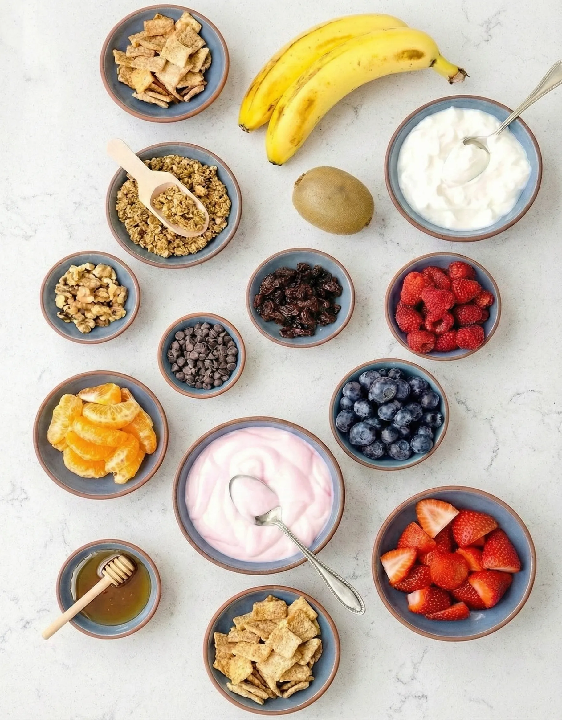 Overhead view of a vibrant yogurt parfait bar setup featuring bowls of white and pink yogurt surrounded by fresh fruit toppings, granola, chocolate chips, nuts, and a sweet honey drizzle.