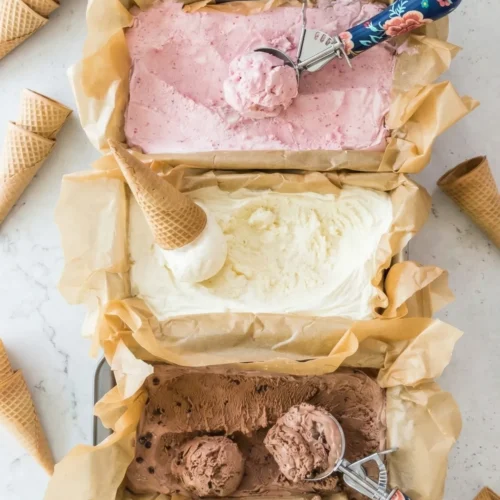 Top-down view of three parchment-lined loaf pans filled with homemade strawberry, vanilla, and chocolate no-churn ice cream, surrounded by waffle cones and floral ice cream scoops on a light countertop.