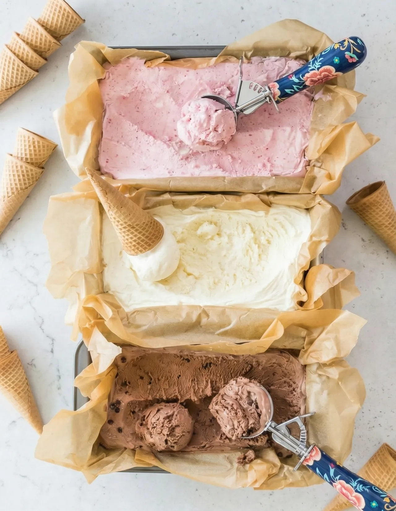no-churn ice cream Top-down view of three parchment-lined loaf pans filled with homemade strawberry, vanilla, and chocolate no-churn ice cream, surrounded by waffle cones and floral ice cream scoops on a light countertop.
