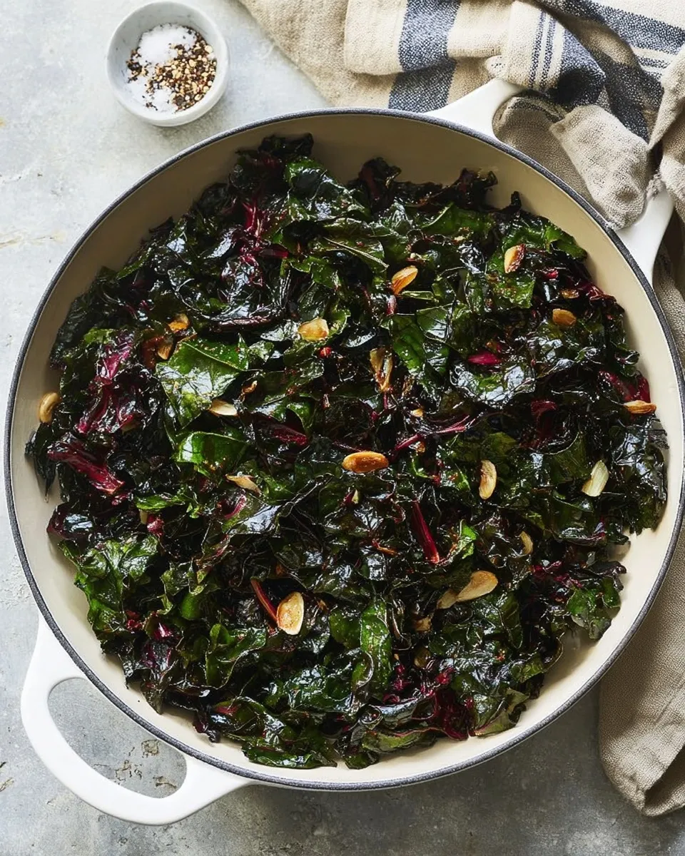 Overhead view of a large white skillet filled with freshly cooked garlic sautéed greens. The dark, glossy leaves and reddish stems are mixed with golden toasted garlic slices, sitting on a grey countertop next to a striped kitchen towel and a small dish of salt and red pepper flakes.