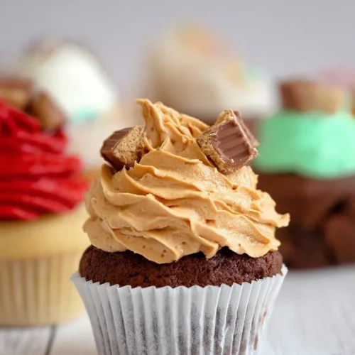 A close-up of a rich chocolate cupcake topped with a tall swirl of fluffy peanut butter buttercream and chopped peanut butter cups, showcasing one of the delicious customizable cupcakes with frosting options on a white wooden surface.