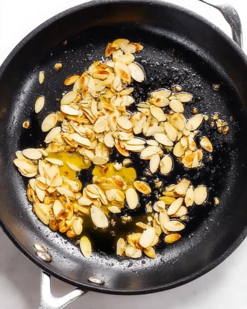 Sliced almonds toasting to a golden brown in a black skillet with oil for green beans almondine.