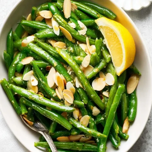 A white serving bowl filled with vibrant green beans with almonds, garnished with toasted sliced almonds, a fresh lemon wedge, and a silver serving spoon.