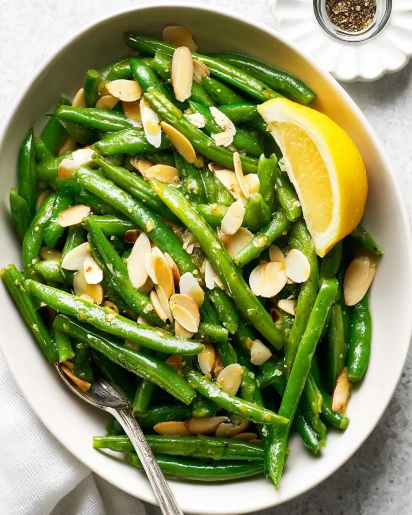 A white serving bowl filled with vibrant green beans with almonds, garnished with toasted sliced almonds, a fresh lemon wedge, and a silver serving spoon.