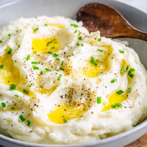 A close-up view of a bowl filled with fluffy mashed potatoes featuring deep swirls holding pools of melted butter, garnished with freshly chopped green chives and cracked black pepper, with a rustic wooden spoon resting on the right side.