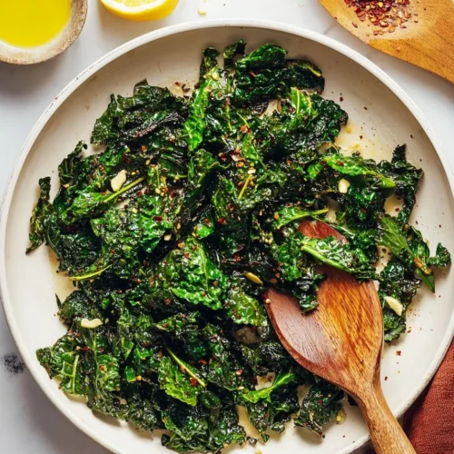 Overhead view of cooked garlic sautéed kale in a white skillet, featuring glossy green leaves topped with golden garlic slices and red pepper flakes. A wooden spoon rests in the pan, with a fresh lemon half, a small dish of olive oil, and extra spices arranged beautifully on the white surface nearby.
