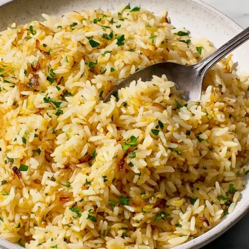 A close-up shot of fluffy, golden easy rice pilaf in a textured ceramic bowl, speckled with fresh green parsley and toasted orzo, with a silver spoon lifting a perfect bite.