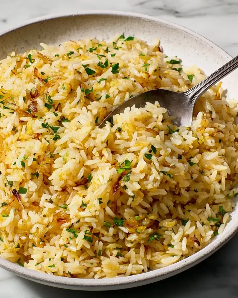 A close-up shot of fluffy, golden easy rice pilaf in a textured ceramic bowl, speckled with fresh green parsley and toasted orzo, with a silver spoon lifting a perfect bite.