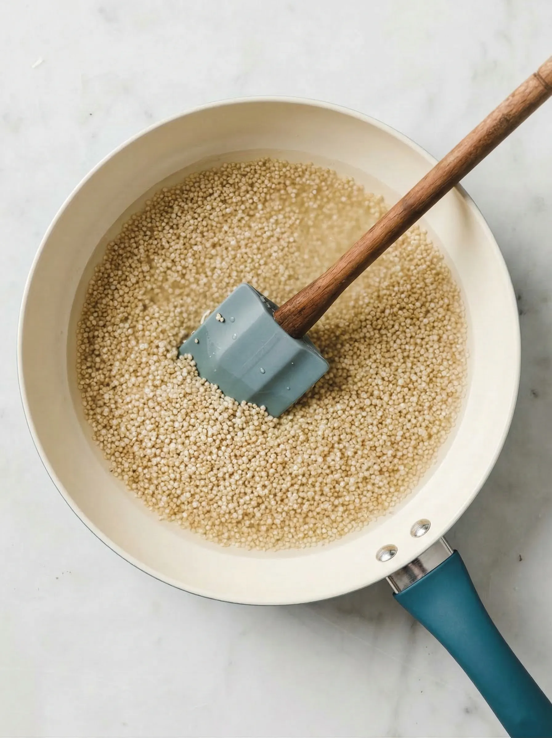 Overhead view of uncooked quinoa and water in a white saucepan with a teal handle, sitting on a marble countertop. A grey silicone spatula with a wooden handle rests inside the pot, ready to stir for an easy Mediterranean grain salad.