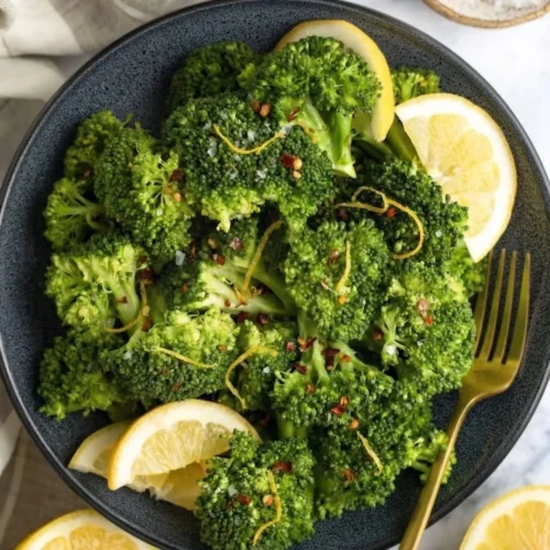 An overhead shot of vibrant green steamed broccoli in a dark ceramic bowl, garnished with fresh lemon slices, bright yellow zest, and red chili flakes. A gold fork rests on the edge of the bowl alongside fresh lemon halves and a small dish of salt on a light marble background, showcasing a perfectly seasoned quick vegetable side dish.