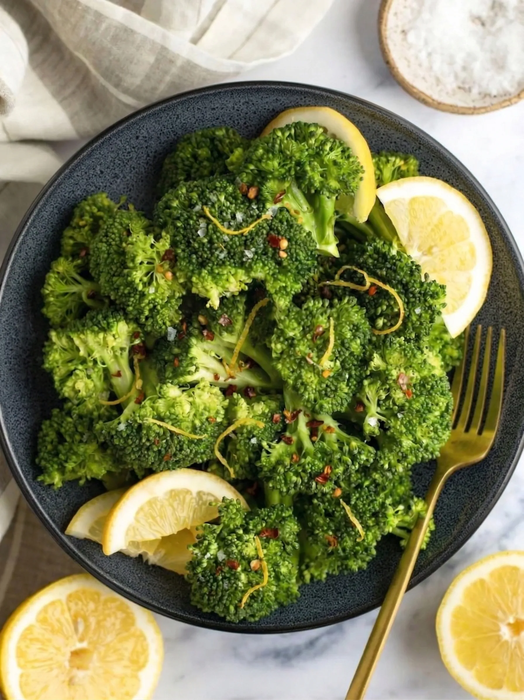 An overhead shot of vibrant green steamed broccoli in a dark ceramic bowl, garnished with fresh lemon slices, bright yellow zest, and red chili flakes. A gold fork rests on the edge of the bowl alongside fresh lemon halves and a small dish of salt on a light marble background, showcasing a perfectly seasoned quick vegetable side dish.