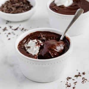 Close-up of a white ramekin filled with thick, glossy homemade chocolate pudding recipe. A silver spoon rests inside the dish, scooping up the pudding to reveal its silky texture. The dessert is topped with a dollop of whipped cream and dark chocolate shavings, set against a bright white marble background.