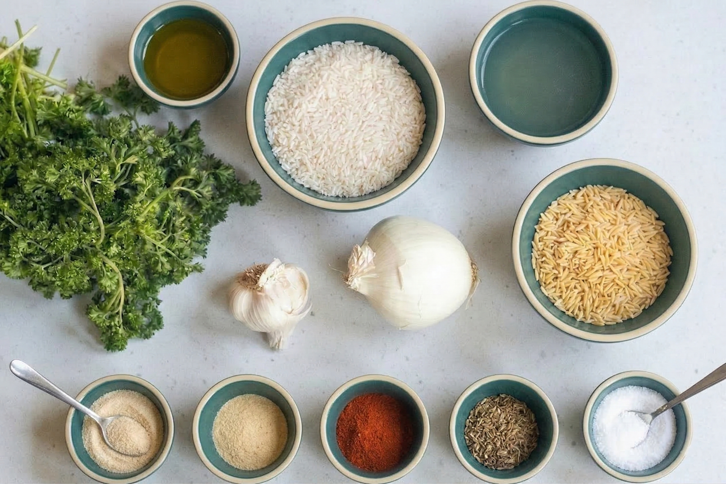 Overhead flat lay of easy rice pilaf ingredients on a light countertop. Matching teal bowls hold long grain white rice, orzo pasta, olive oil, broth, and five different dry spices, all arranged around a bunch of fresh parsley, a whole white onion, and a head of garlic.