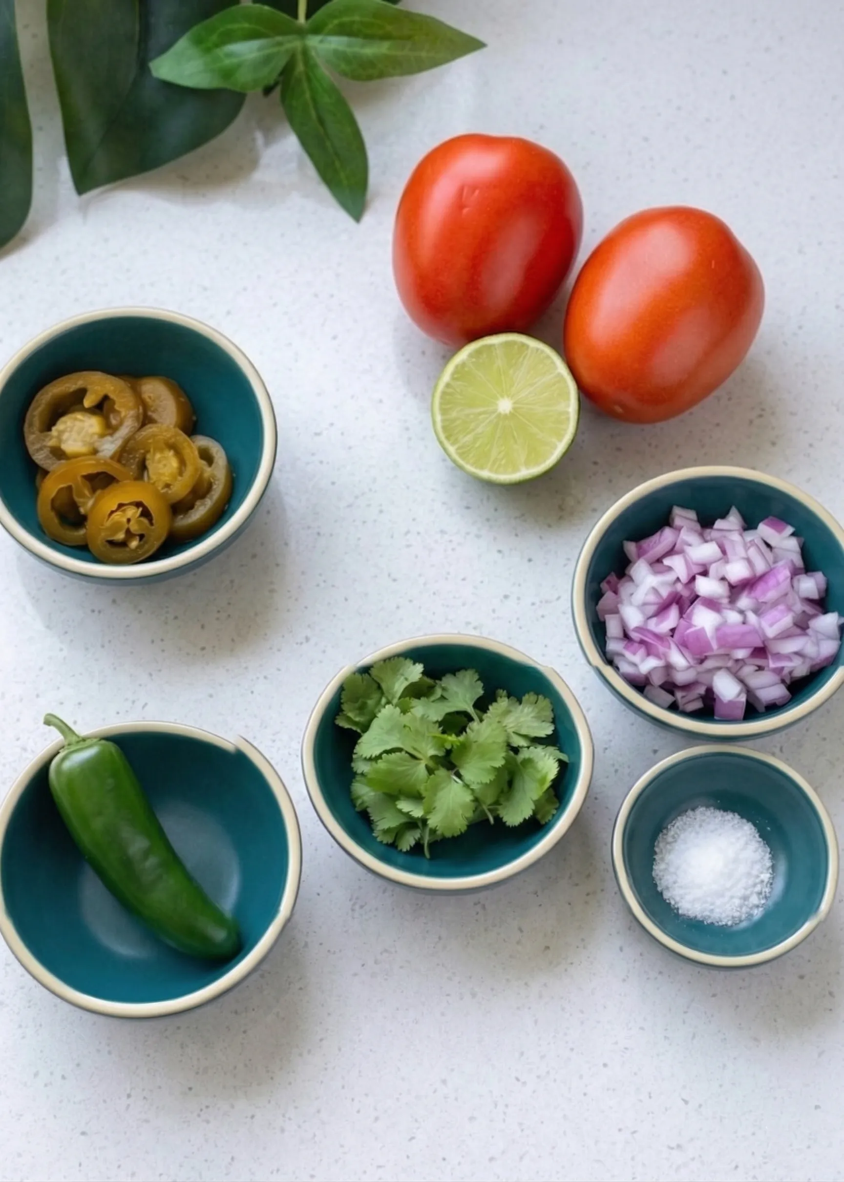 Overhead view of fresh pico de gallo ingredients arranged on a white speckled countertop, including whole Roma tomatoes, half a lime, and small teal bowls filled with diced red onion, fresh cilantro, a whole jalapeño, pickled jalapeño slices, and coarse salt.