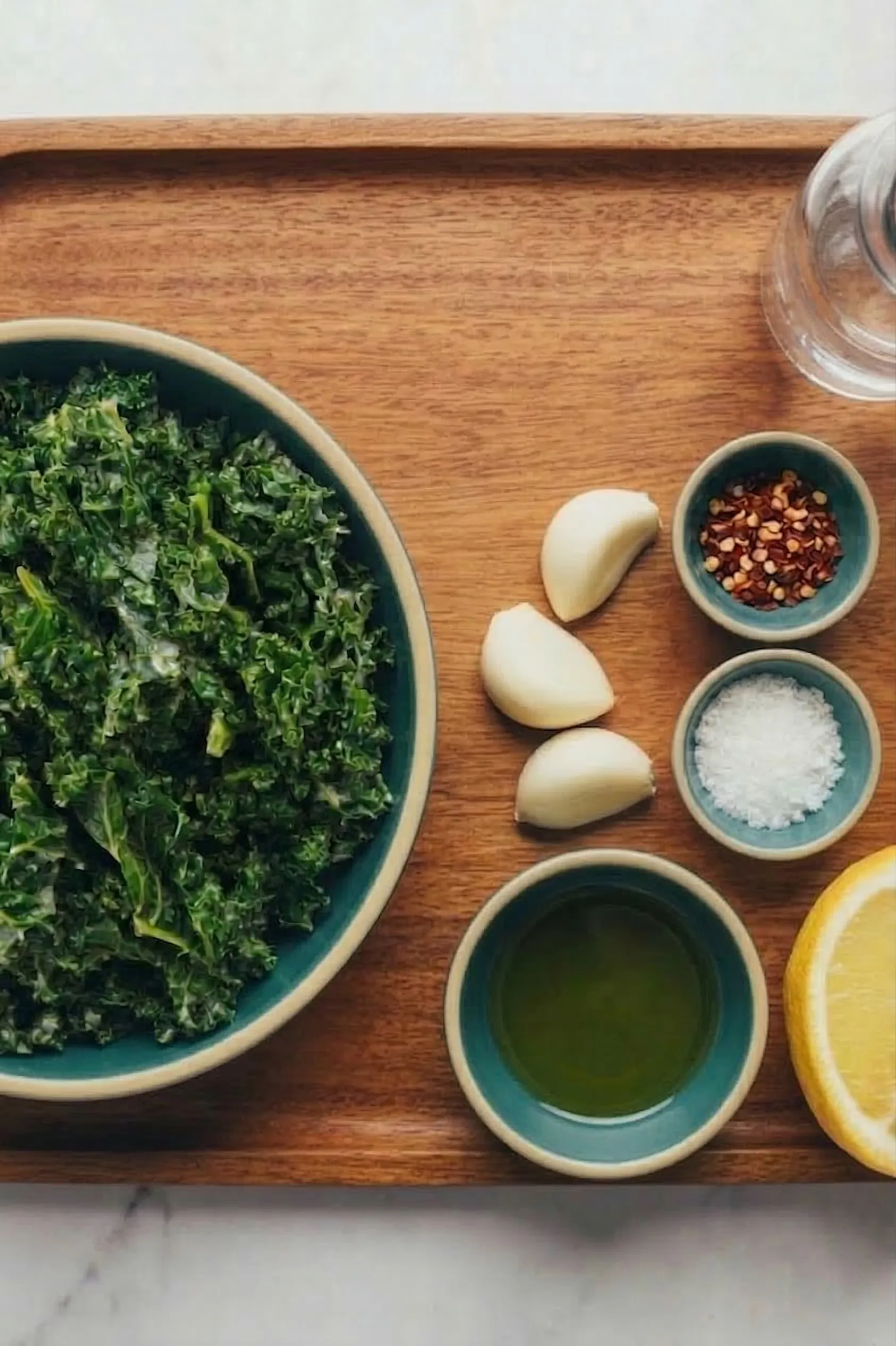 Overhead view of fresh ingredients for garlic sautéed greens on a wooden tray, featuring a large bowl of chopped kale, fresh garlic cloves, olive oil, red pepper flakes, sea salt, and a lemon half.