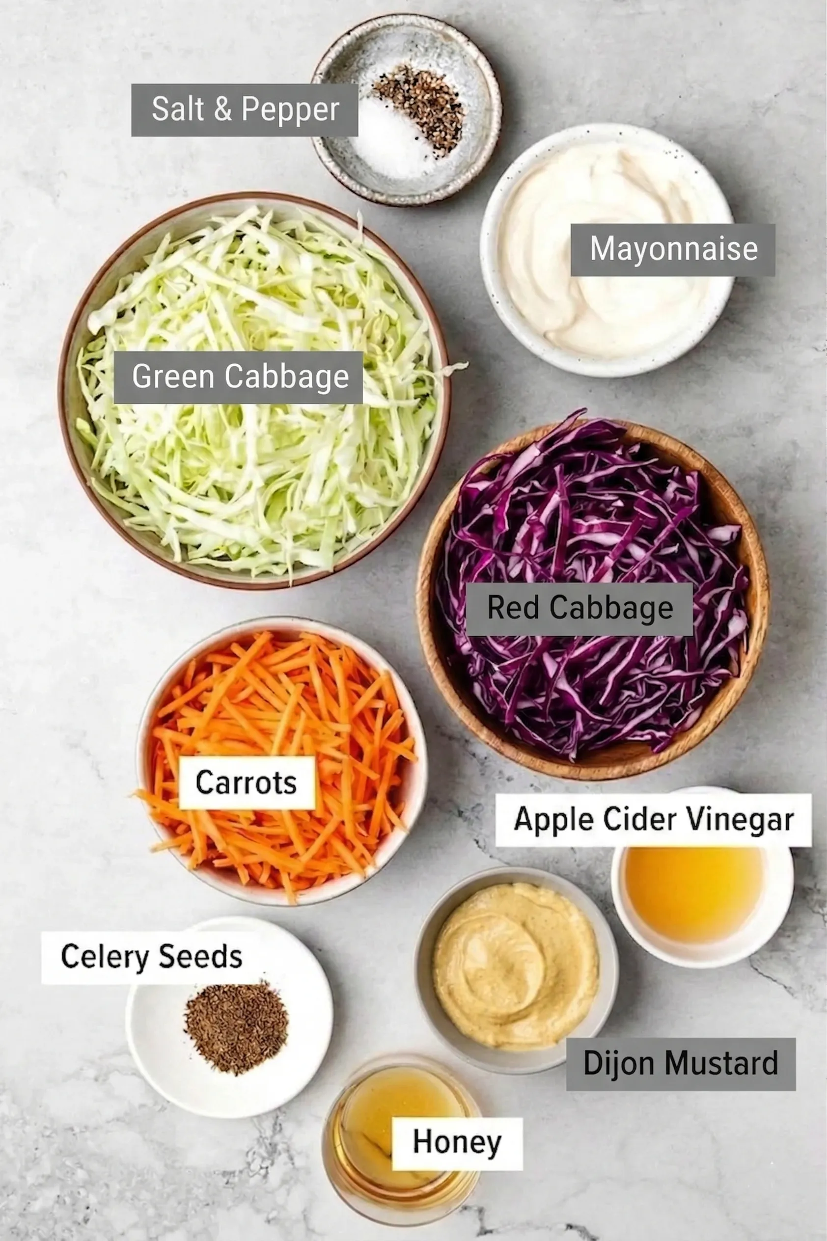 Overhead view of fresh, vibrant ingredients for a creamy coleslaw dressing with apple cider vinegar, featuring shredded green and red cabbage, carrots, mayonnaise, honey, Dijon mustard, celery seeds, and salt and pepper in small bowls.