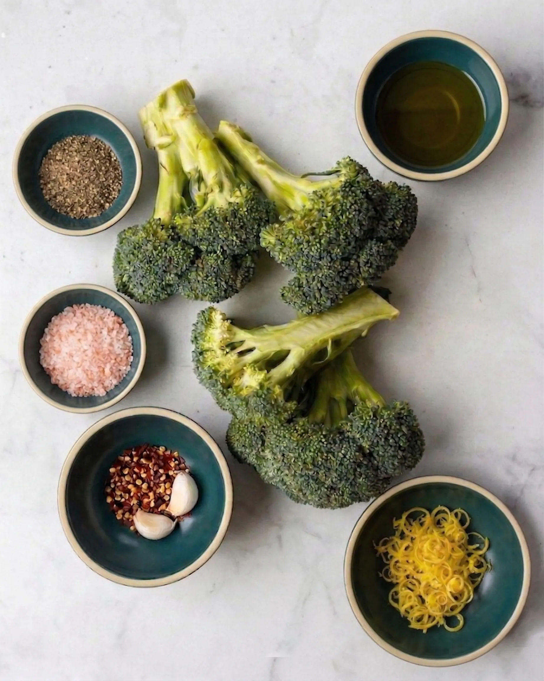 An overhead shot of fresh ingredients for quick steamed broccoli arranged on a light marble countertop. Bright green heads of fresh broccoli sit in the center, surrounded by small teal bowls containing coarse pink salt, black pepper, red chili flakes with garlic cloves, fresh bright yellow lemon zest, and extra virgin olive oil.