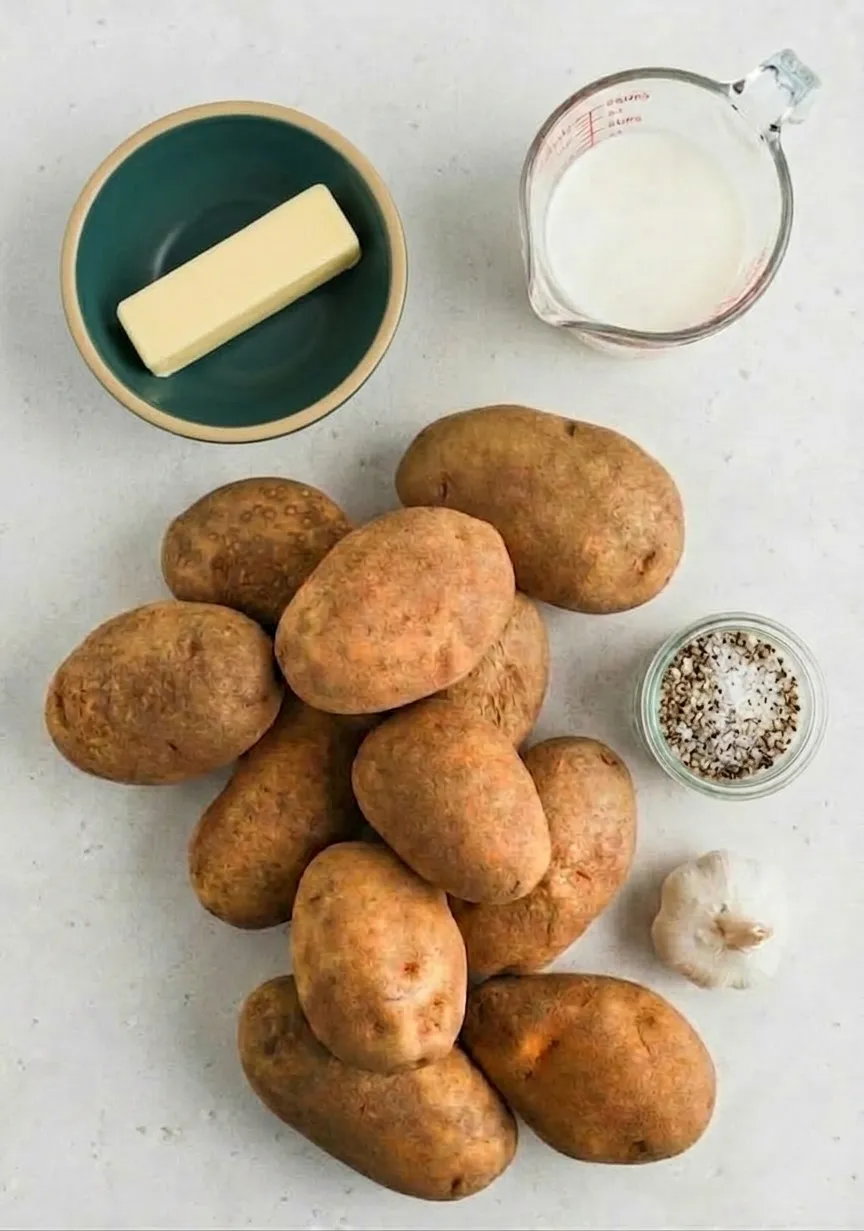 Overhead view of simple mashed potato ingredients on a light countertop, including a pile of whole potatoes, a stick of butter in a green bowl, a glass measuring cup of milk, a small jar of mixed salt and pepper, and a head of garlic