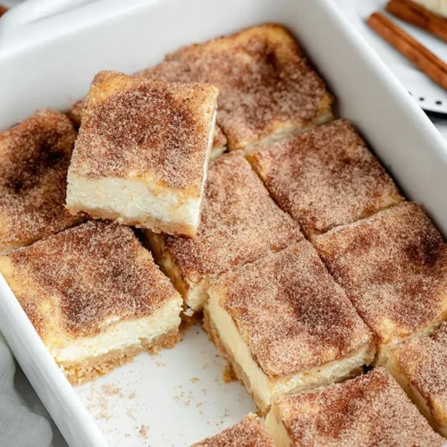 A white baking dish filled with pre-cut churro cheesecake bars, highlighting the thick cream cheese center and cinnamon-sugar crust on a lifted slice.