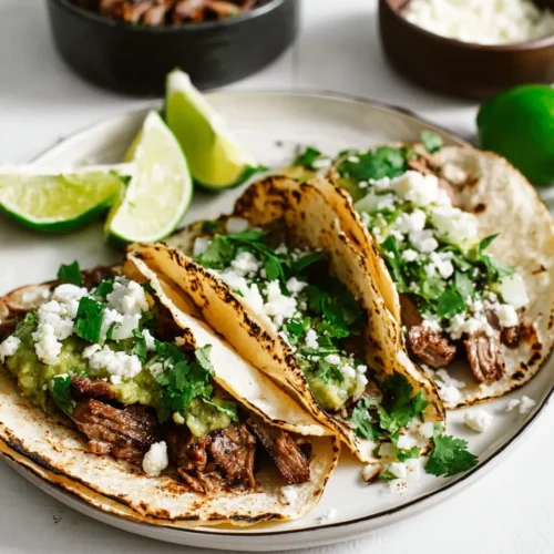 A close-up shot of three authentic carne asada tacos with corn tortillas, filled with tender grilled steak, mashed avocado, crumbled cotija cheese, diced onions, and fresh cilantro, plated alongside fresh lime wedges.