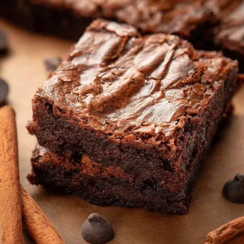 A close-up shot of a thick, fudgy Mexican spiced chocolate brownie resting on brown parchment paper. The brownie features a shiny, crinkly top crust and is surrounded by whole cinnamon sticks and scattered semi-sweet chocolate chips.