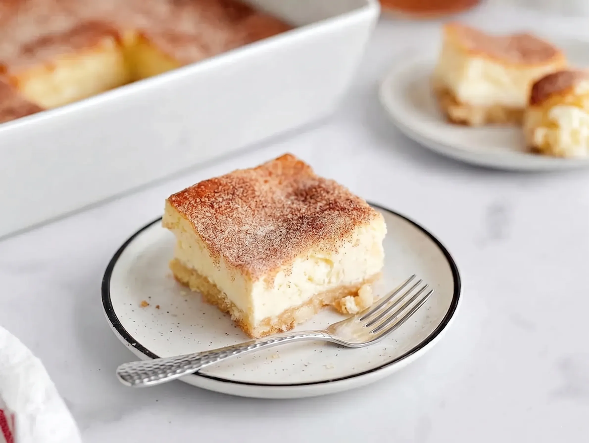 A close-up of a single churro cheesecake bar served on a small plate, showing a thick cream cheese layer and a flaky, cinnamon-dusted crust.