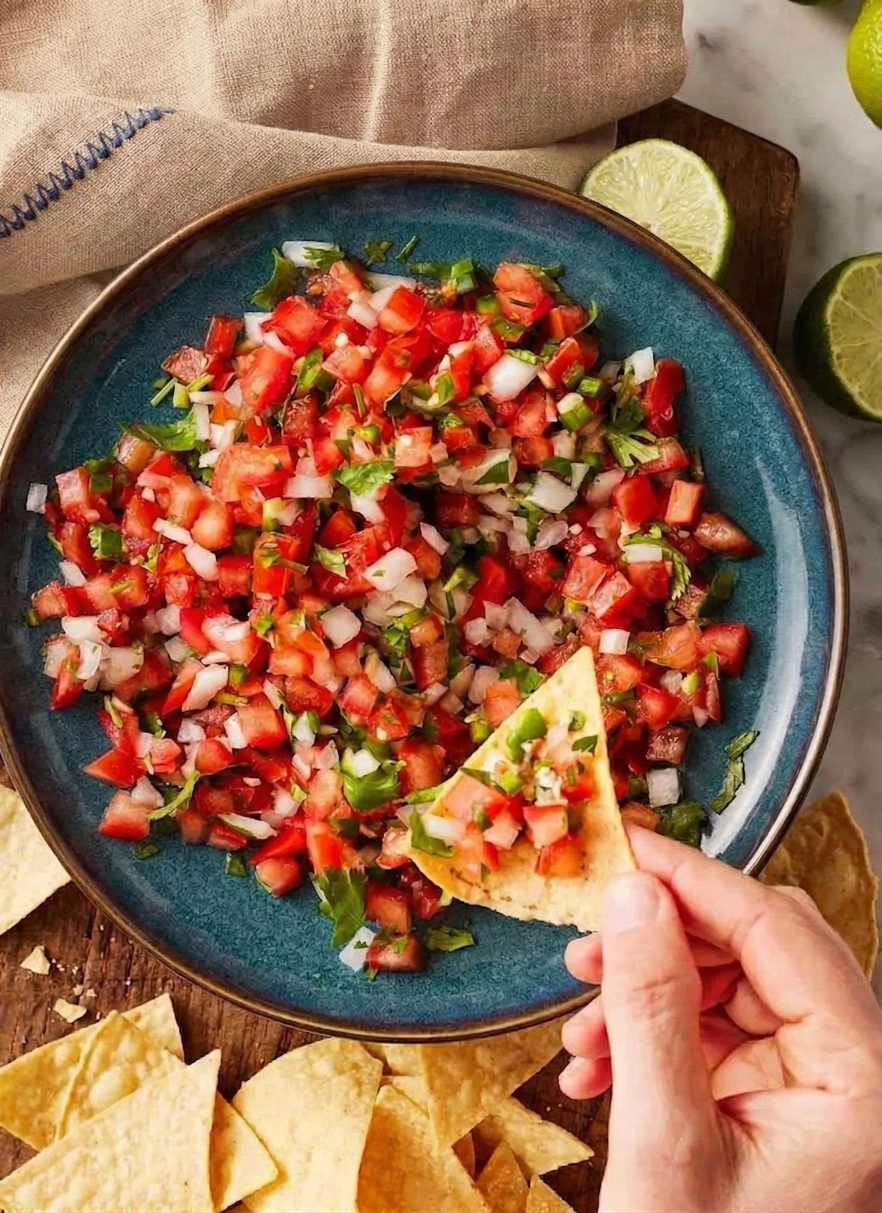 Top-down view of a hand using a yellow corn tortilla chip to scoop fresh pico de gallo from a rustic blue bowl. The salsa is a chunky mix of diced red tomatoes, white onion, green jalapeño, and cilantro. The bowl rests on a wooden surface alongside fresh lime halves, scattered tortilla chips, and a tan linen napkin.