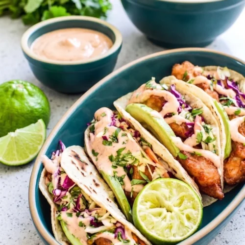 Alt Text A close-up of four assembled Baja fish tacos in a teal oval serving dish, topped with fresh avocado slices, purple and green cabbage slaw, and a drizzle of pink chipotle mayo. Small teal bowls of extra slaw and sauce, along with fresh limes and cilantro, sit in the background on a bright, speckled countertop.