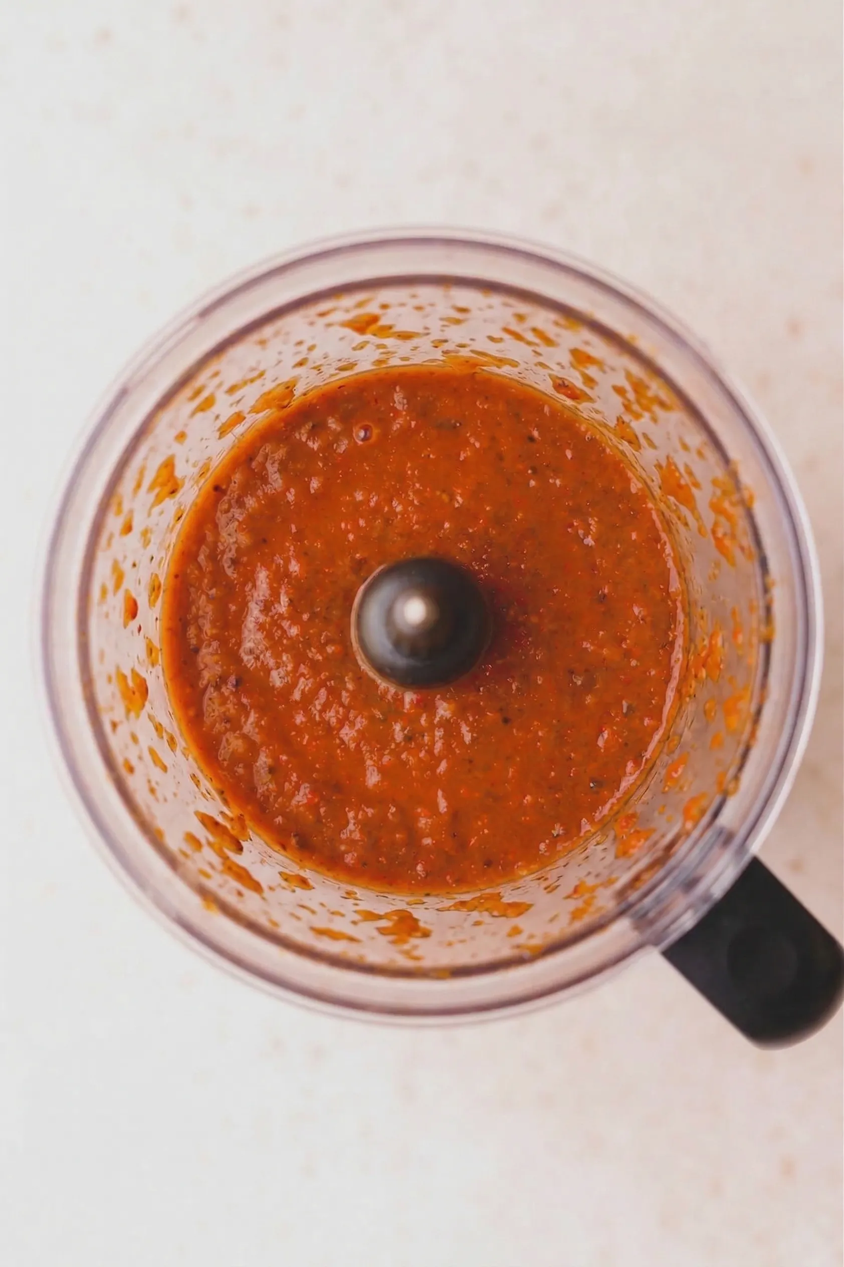 An overhead view looking down into a clear blender pitcher filled with a thick, brightly colored reddish-orange blended adobo sauce.