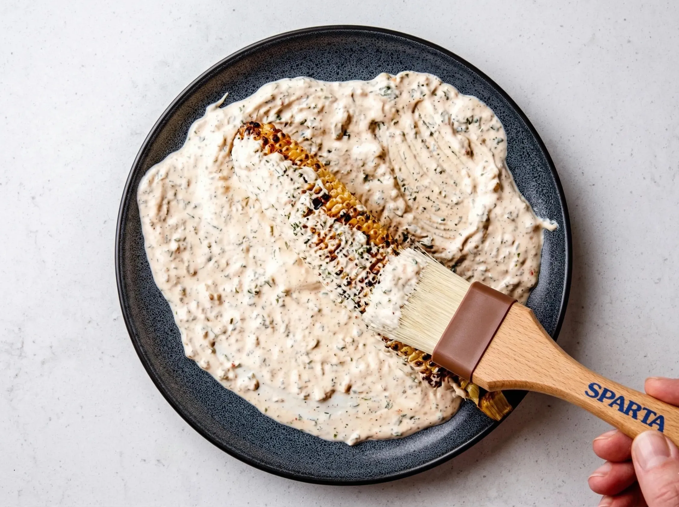 A close-up overhead shot of a hand using a wooden pastry brush to generously coat a charred ear of corn with a creamy, herb-speckled mayo sauce on a dark plate.