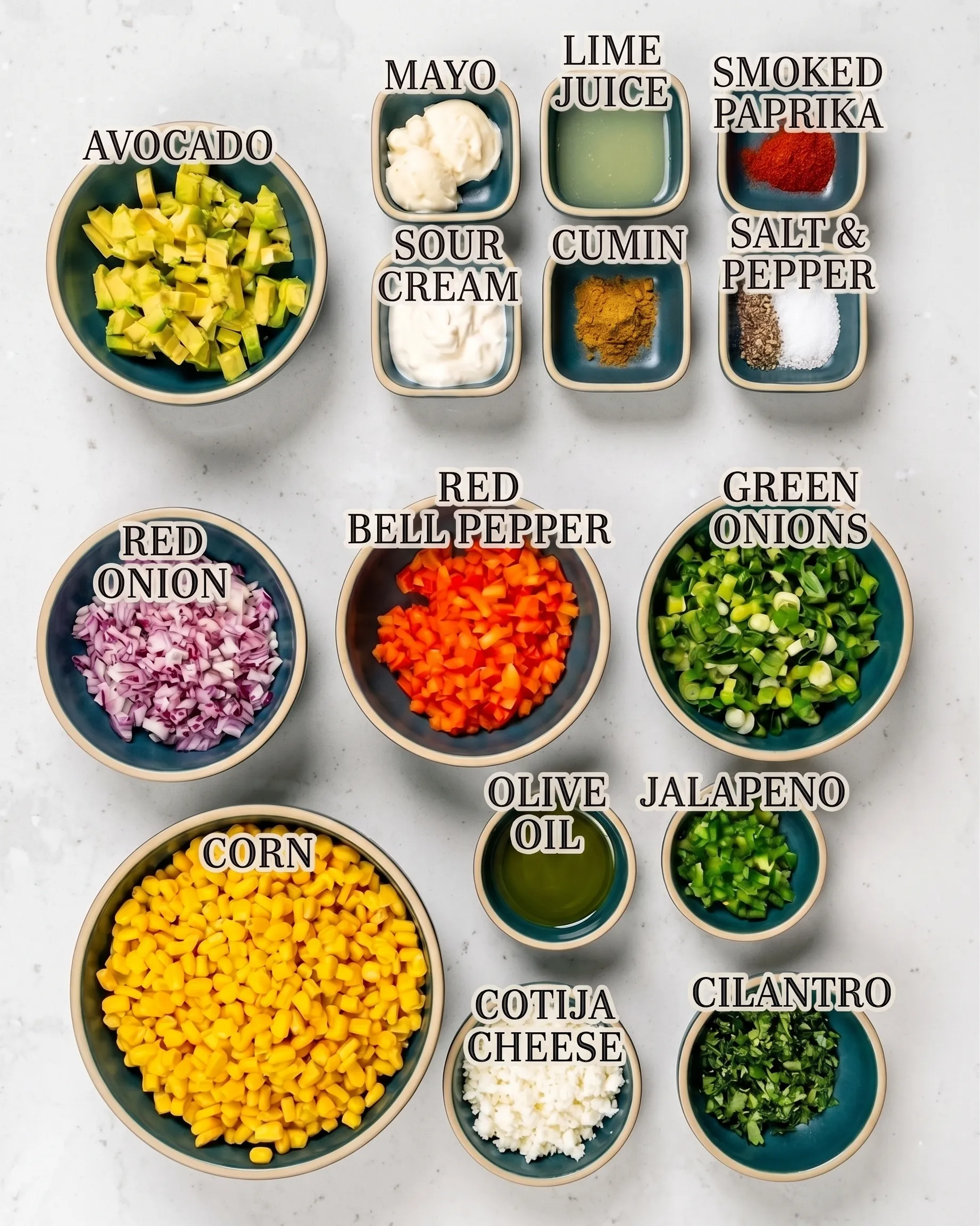 A top-down view of neatly arranged teal bowls on a light, speckled countertop, filled with ingredients for Mexican street corn salad. Text overlays identify each item: diced avocado, diced red onion, corn kernels, mayo, sour cream, diced red bell pepper, olive oil, crumbled cotija cheese, lime juice, cumin, sliced green onions, diced jalapeno, chopped cilantro, smoked paprika, and salt & pepper.