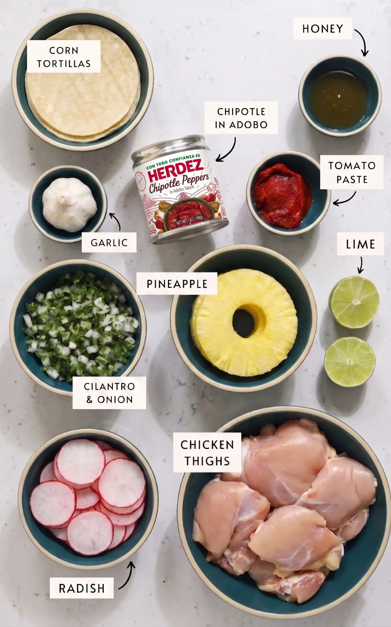 Overhead view of labeled ingredients for chicken tacos al pastor arranged in teal bowls on a white countertop, including corn tortillas, whole garlic, diced cilantro and onion, sliced radishes, a can of chipotle peppers in adobo, pineapple rings, raw chicken thighs, tomato paste, honey, and lime halves.