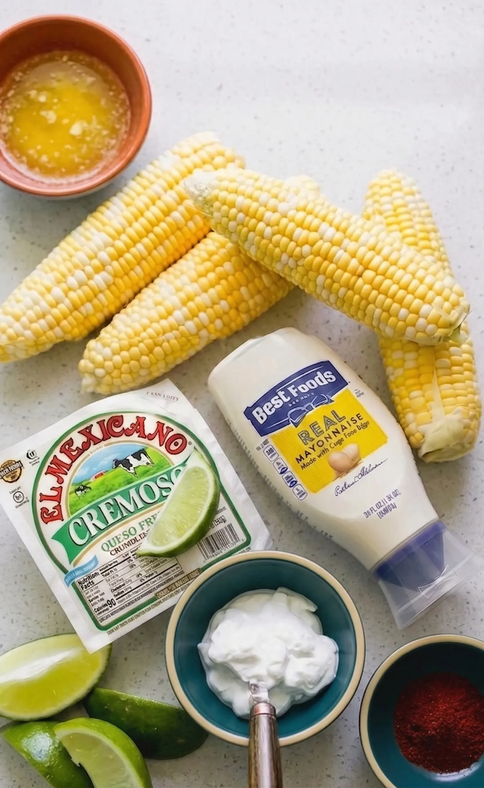 Overhead shot of fresh ingredients for Mexican Street Corn (Elote) laid out on a light speckled countertop, including four husked ears of raw sweet corn, mayonnaise, a package of crumbly queso fresco, small bowls of melted butter, crema, and red chili powder, alongside fresh lime wedges.