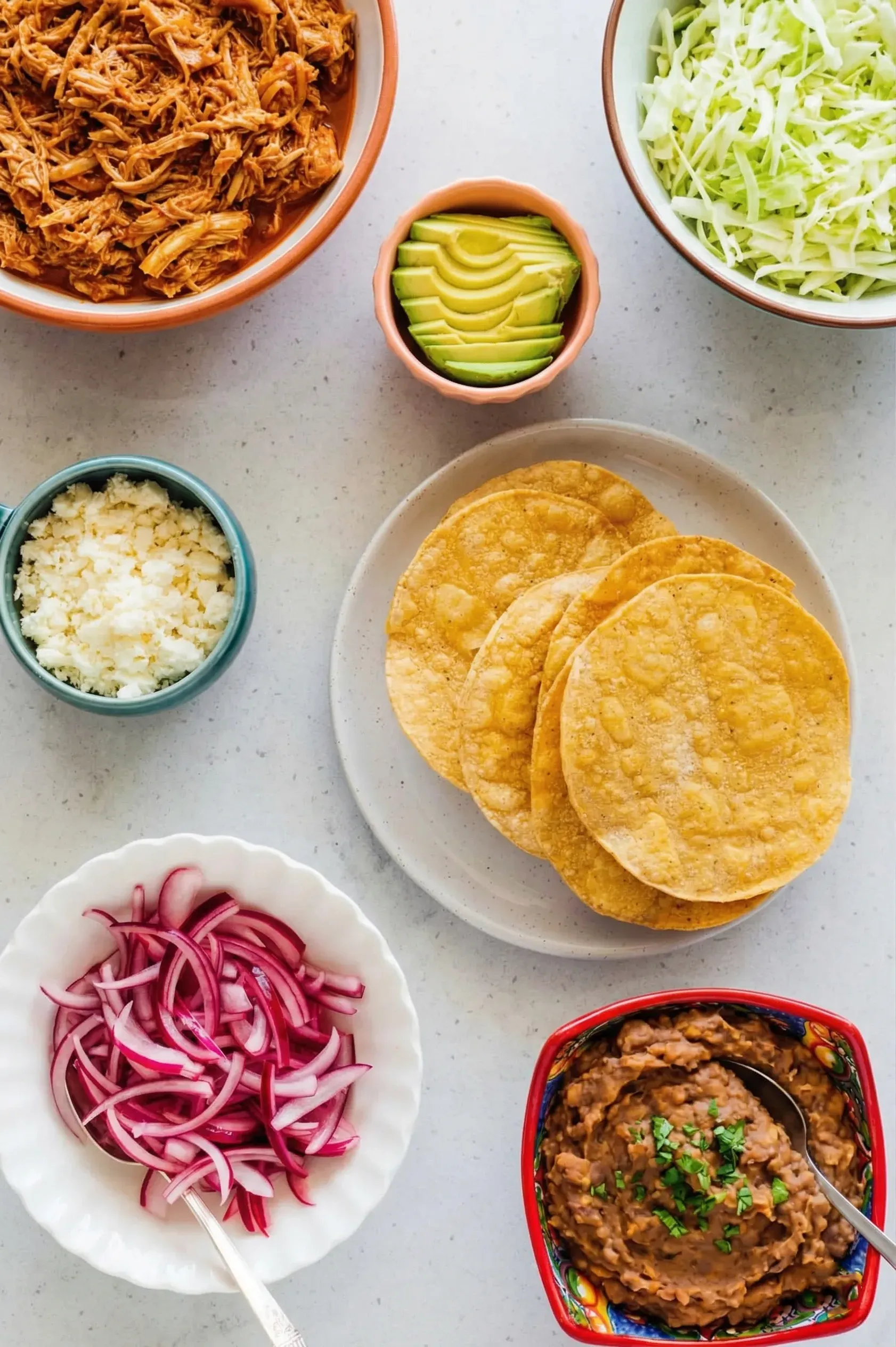 ingredients for chicken tinga tostadas An overhead flat lay of ingredients for chicken tinga tostadas arranged on a light grey speckled countertop. There are individual bowls containing shredded chicken in a red chipotle sauce, shredded iceberg lettuce, sliced avocado, crumbled cotija cheese, bright pink pickled red onions, and refried beans garnished with cilantro, all surrounding a central plate of four crispy, golden tostada shells.