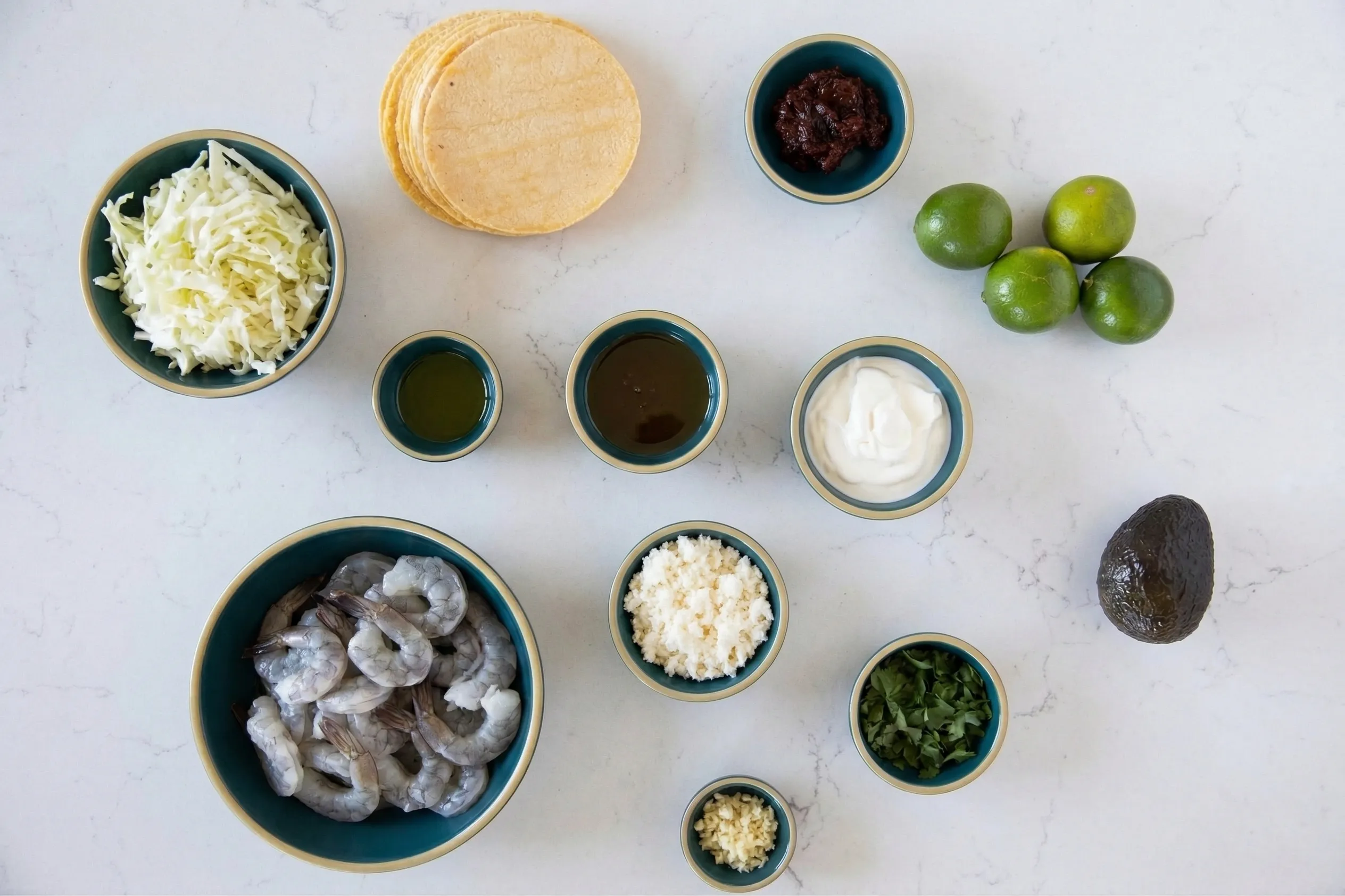 An overhead flat-lay view of shrimp taco ingredients arranged in dark teal bowls on a white marble countertop. The spread includes raw shrimp, shredded cabbage, a stack of corn tortillas, whole limes, an avocado, sour cream, chipotle peppers, crumbled cheese, chopped cilantro, minced garlic, and small bowls of oil and sauce.
