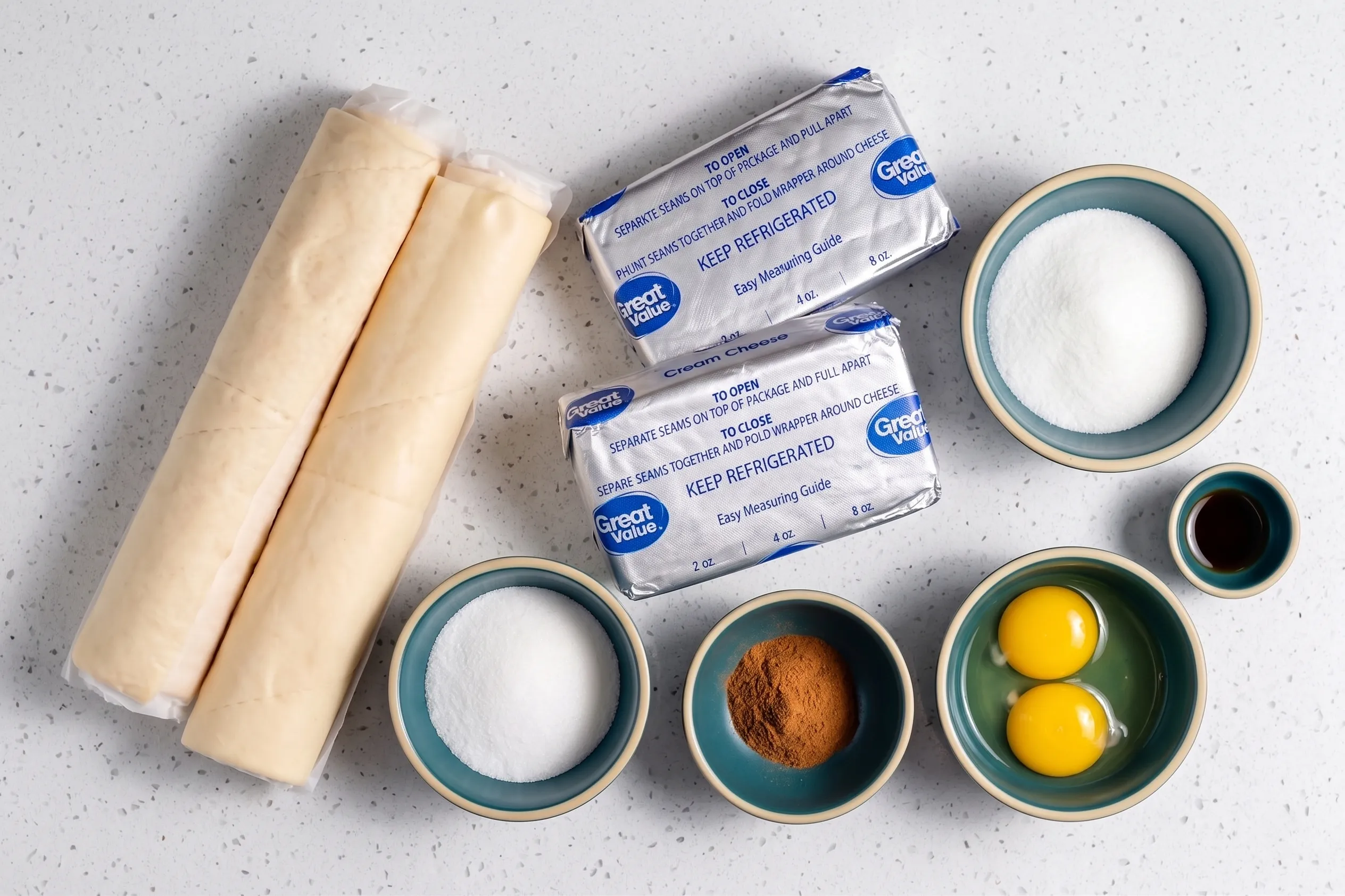 Overhead view of ingredients for churro cheesecake bars, including crescent roll dough, cream cheese, sugar, cinnamon, eggs, and vanilla extract arranged on a white counter.