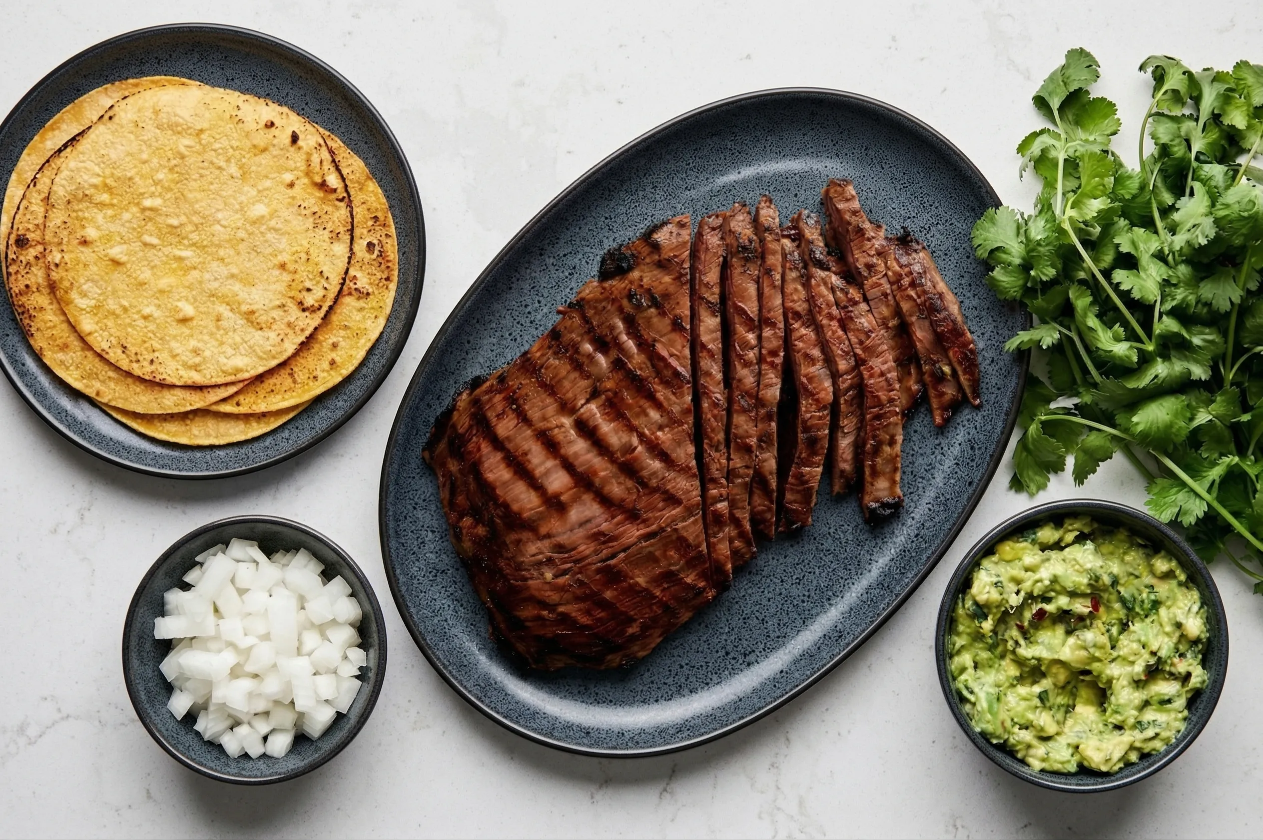 An overhead shot of prep ingredients for authentic carne asada tacos with corn tortillas , featuring a large blue platter of grilled steak demonstrating how to slice carne asada against the grain. The meat is surrounded by small bowls of diced white onion and mashed avocado, alongside fresh cilantro and a stack of warm tortillas.