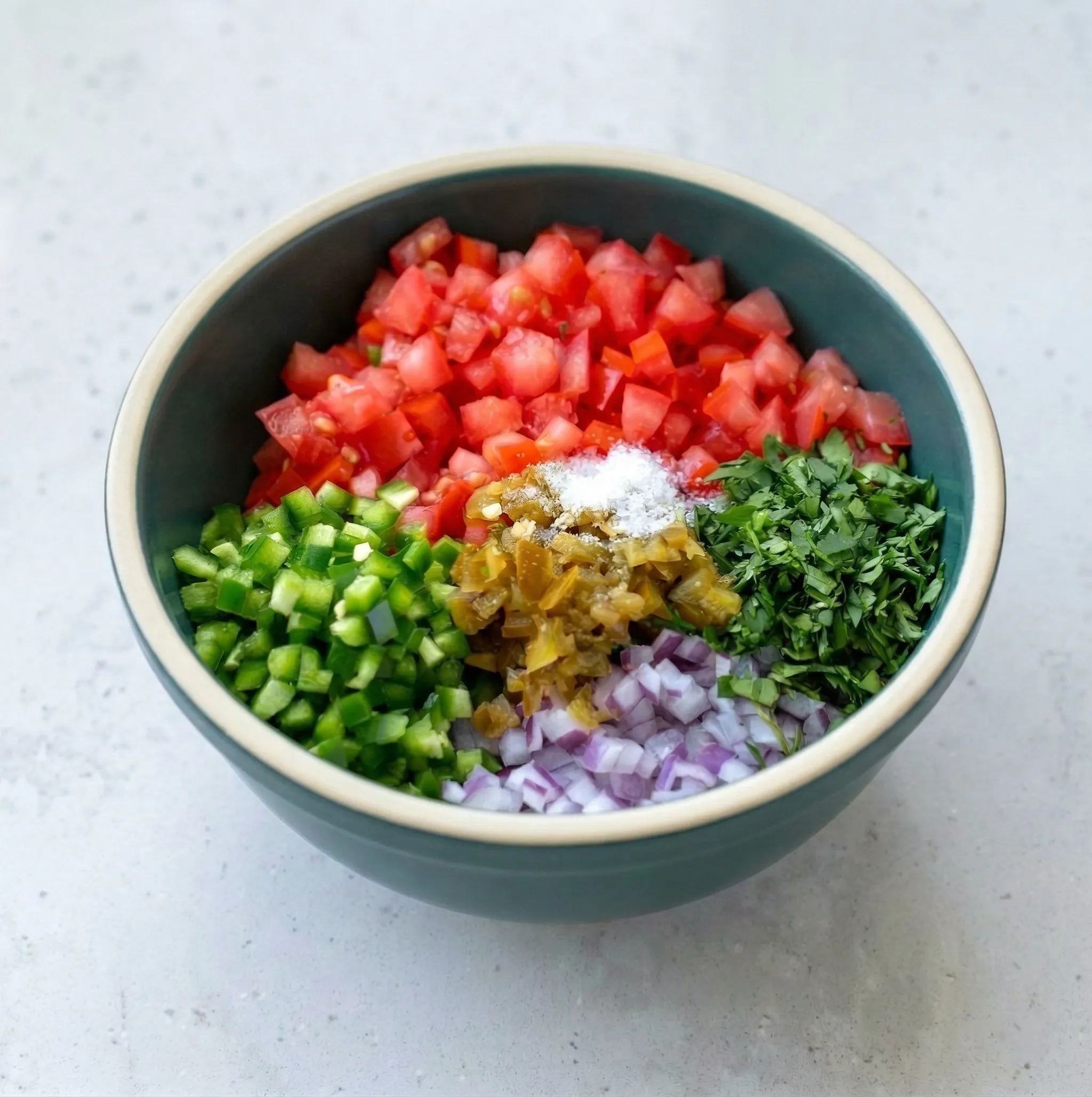 Overhead view of a teal bowl on a white speckled countertop, filled with neatly separated, diced ingredients for pico de gallo: fresh tomatoes, red onion, green jalapeños, chopped cilantro, pickled jalapeños, and coarse salt.