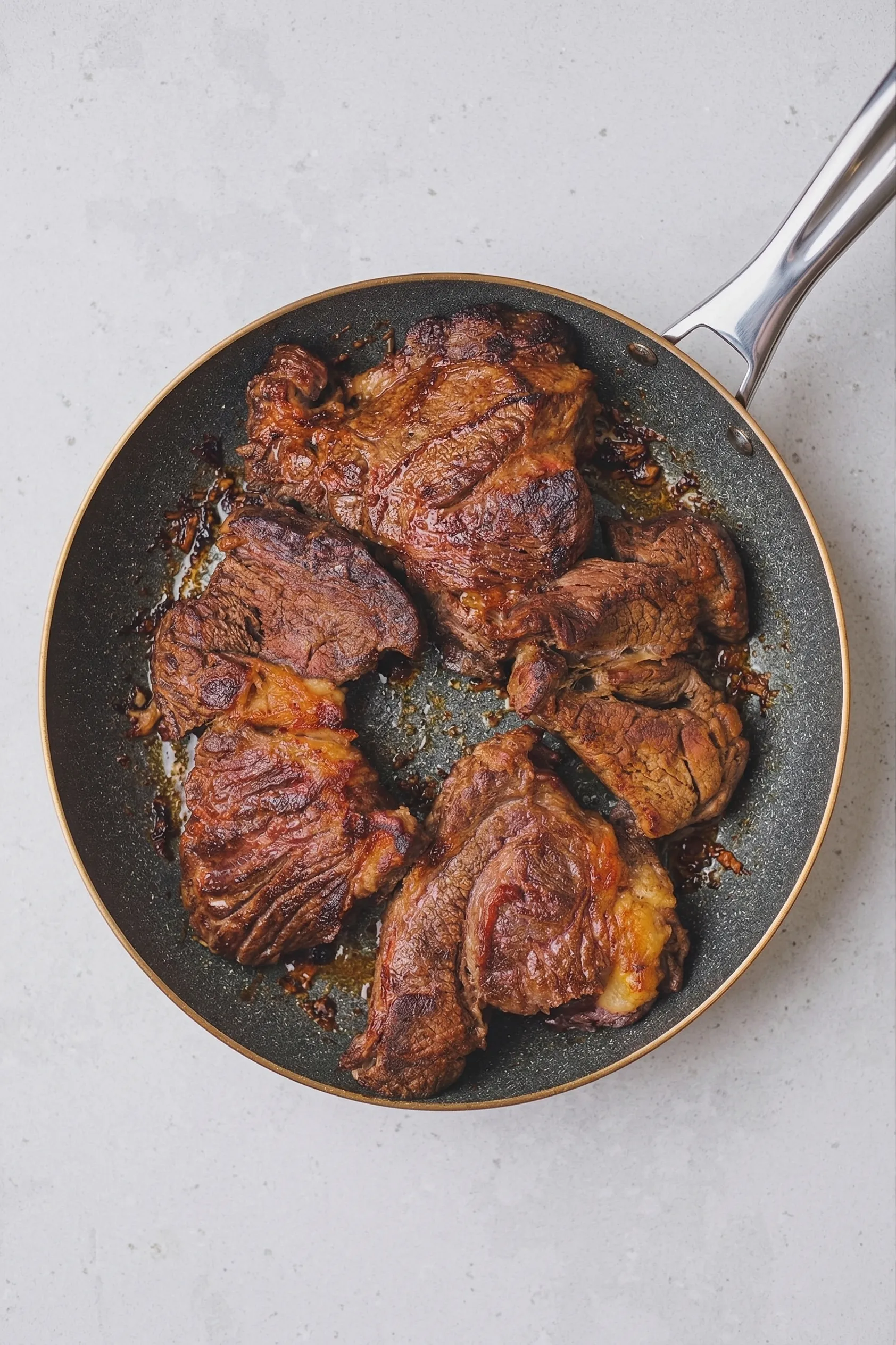 An overhead shot of a dark frying pan sitting on a light countertop, filled with several large chunks of beef chuck roast that have been seared to a deep, caramelized brown.