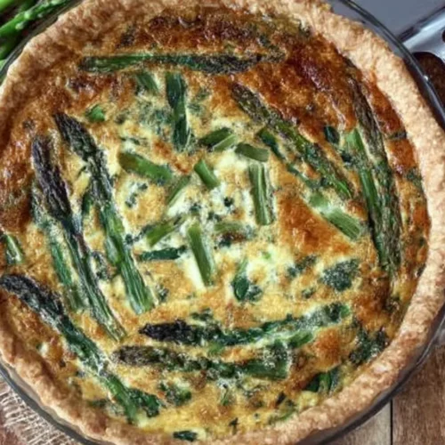An overhead view of a freshly baked, golden-brown asparagus quiche in a glass pie dish, sitting on a rustic wooden table next to raw green asparagus spears and a serving spatula