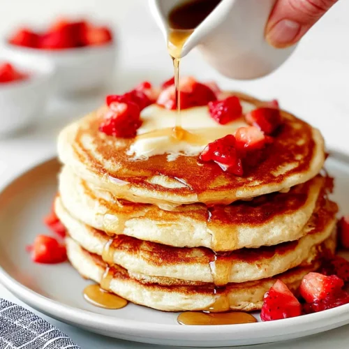 A close-up shot of a hand pouring warm maple syrup over a stack of fluffy ricotta pancakes topped with melting butter and fresh strawberries.