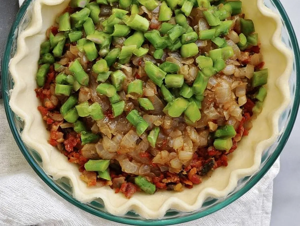 An unbaked pie crust in a round glass pie pan layered with crumbled bacon, golden sautéed diced onions, and bright green chopped asparagus spears.